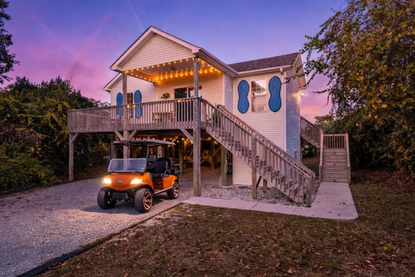 Elevated beach house with white siding, blue oval windows, lit deck, and orange golf cart parked underneath at dusk.