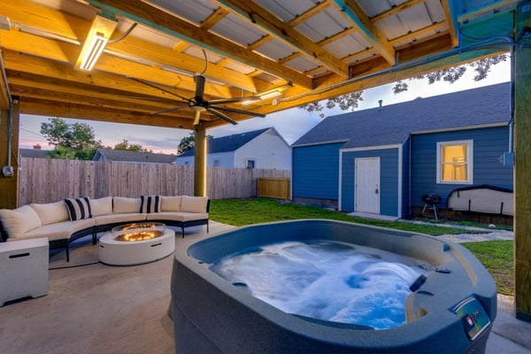 Backyard patio with hot tub, curved sectional sofa, fire pit, wooden pergola, and blue shed at dusk