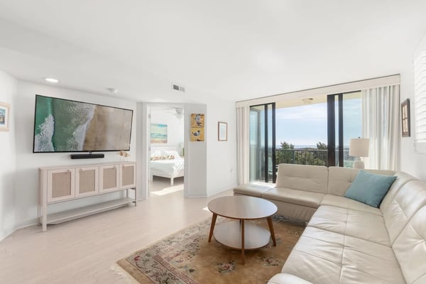 Bright living room with cream sofa, round coffee table, wall-mounted TV, and ocean view through sliding glass doors